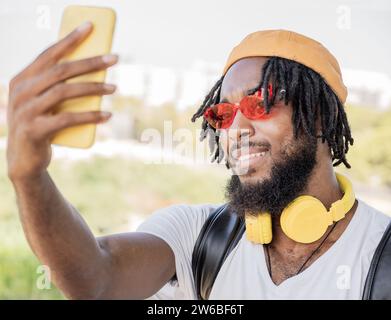 Smiling African American man with dreadlocks wearing red sunglasses and hat with headphones around the neck sitting and taking selfie via smartphone o Stock Photo
