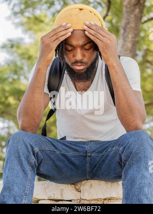 Young African American guy in street style clothes sitting on stone border and touching forehead while having problems listening to sad song in summer Stock Photo