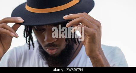 Portrait of young bearded African American guy in white t shirt leaning head forward and adjusting black hat in sunny park Stock Photo