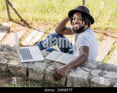 High angle of young African American male blogger in hat sitting on ground touching hat with pen while looking at camera and thinking in park with lap Stock Photo