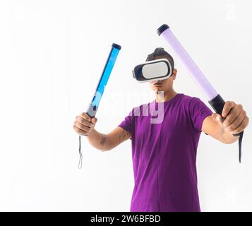 A man in a purple shirt wields two glowing light swords while using a virtual reality headset against a white background Stock Photo