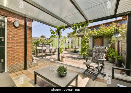 Coffee table with empty chairs in patio under roof of modern house with dining table and plants in backyard on sunny day Stock Photo