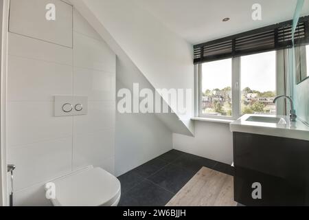 Closed toilet bowl near sink with faucet by window with blinds in white loft bathroom in home Stock Photo