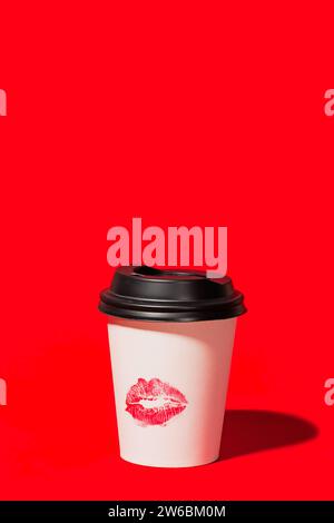 A single white disposable coffee cup with a black lid and a red lipstick kiss mark stands on a vivid red background, creating a bold and romantic stat Stock Photo