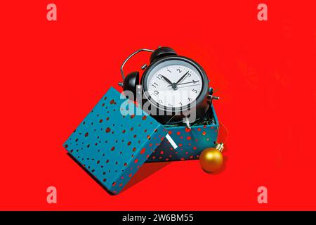 A black alarm clock with white headphones and a teal gift box with polka dots, decorated with a gold Christmas ornament on a vibrant red background. Stock Photo