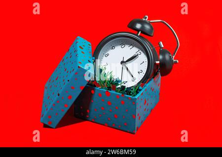 A black vintage alarm clock nestled in an open blue polka-dotted gift box against a red background. Stock Photo