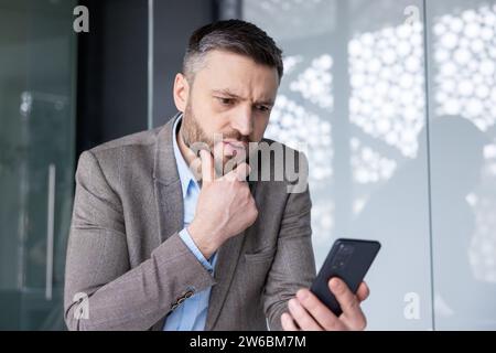 Upset frustrated man holding phone inside office, sad businessman reading bad news online, boss experienced financier accountant unhappy with workplace achievement results. Stock Photo