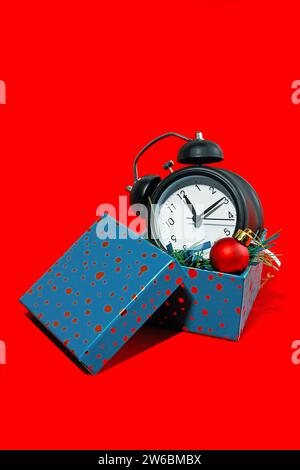 Black alarm clock and red Christmas bauble emerging from a blue gift box set against a vibrant red background. Stock Photo