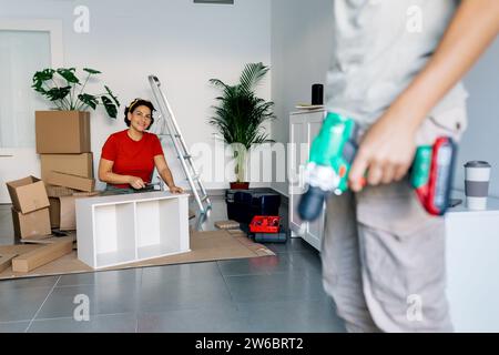 Positive woman in red t shirt looking at anonymous son with drill machine while assembling small shelf with hammer during relocation into new modern a Stock Photo