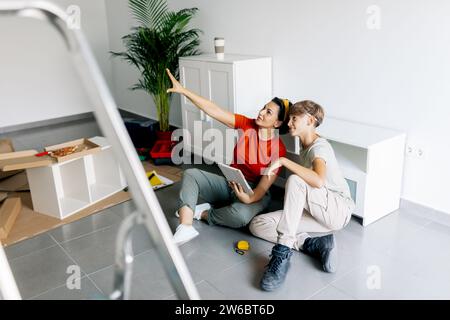 High angle side view of mom sitting together with son holding notepad and pointing upwards during moving in new apartment Stock Photo