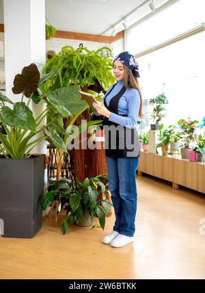 Full body of young female florist holding tablet and checking leaves of potted plant while working in floral shop Stock Photo