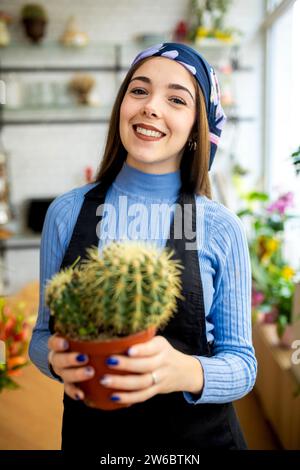 Positive young female gardener wearing headscarf and apron standing in flower shop and holding potted cactus during work Stock Photo