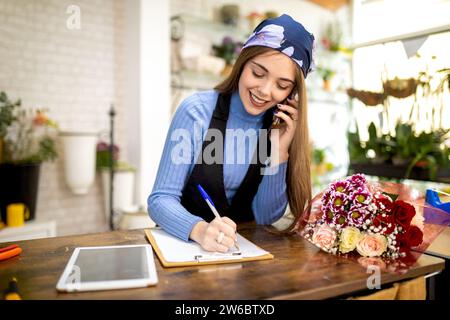 Positing young female florist in casual clothes and bandana talking on smartphone while writing down order at wooden desk in flower shop Stock Photo