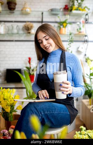 Positive young female florist holding disposable coffee cup while using digital table sitting on stool and working in flower shop Stock Photo