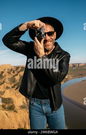 A trendy happy man wearing a black hat and leather jacket taking pictures with a vintage camera against a natural backdrop Stock Photo