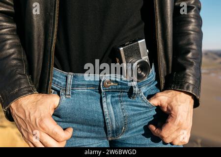 Close-up of a vintage camera tucked into the denim jeans of a man wearing a black leather jacket, no face shown Stock Photo