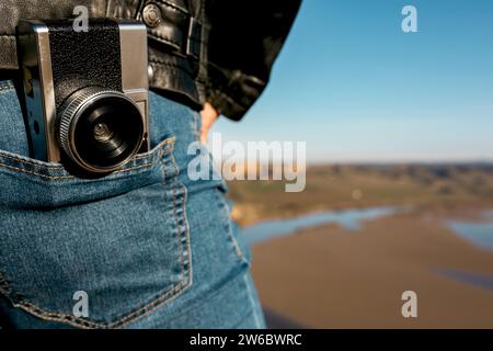 Close-up of a vintage camera tucked into adventurous photographer's jeans, with a river landscape in the background, depicting a sense of adventure an Stock Photo