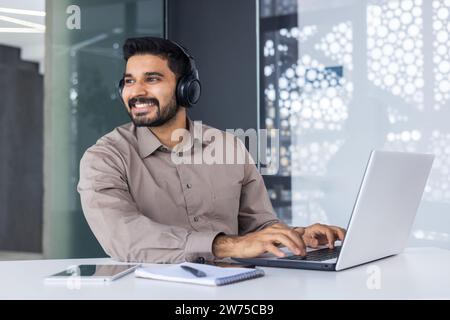 Smiling young man indian programmer and developer working in headphones on laptop, sitting at desk in office and looking away. Stock Photo