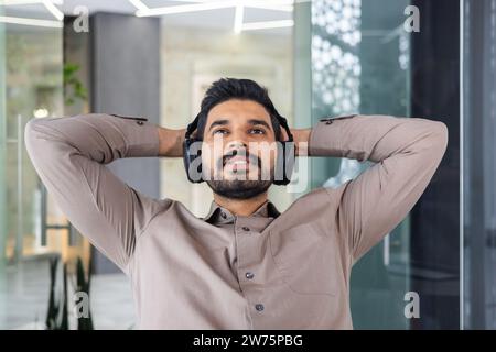 Close-up photo of a young Indian man in headphones who works in the office, sits thoughtfully at the table and rests with his hands behind his head. Stock Photo