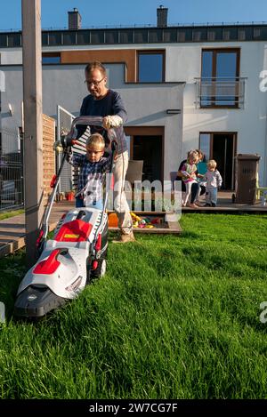 15.09.2018, Poland, Wrzesnia, Wielkopolska - Father and son mowing the lawn in the garden of their terraced house, mother with siblings in the backgro Stock Photo