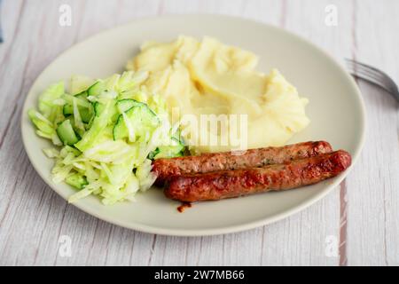 Classic meal: Sausages, mashed potatoes, and cabbage salad with cucumbers on a white plate against a wooden background Stock Photo