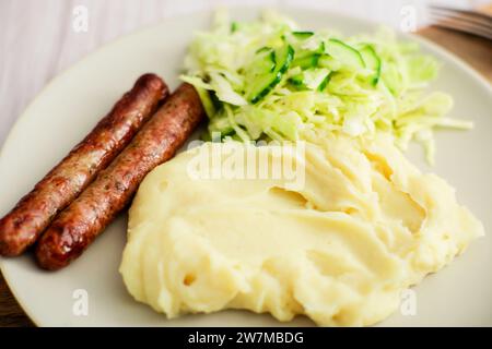 Sausages, mashed potatoes, and a zingy cabbage-cucumber salad elegantly displayed on white dinnerware against a wooden background Stock Photo