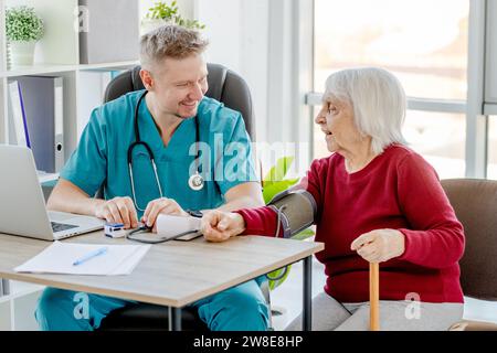 Young, Handsome Doctor Measures Blood Pressure Of Elderly Lady In His Medical Office Stock Photo