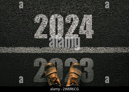 Man Walks On Asphalt And Crosses The Line By 2024. New Year 2024. Men's Legs With Leather Shoes Go To 2024. End Of 2023 And Beginning Of 2024 Stock Photo