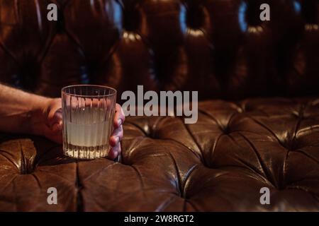 man's hand holding crystal glass with a cocktail on brown Chesterfield sofa couch Stock Photo