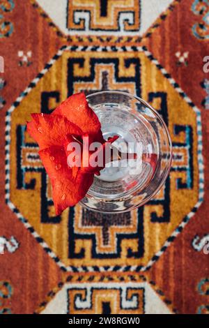 top down, directly above shot of glass of water with red flower on orange, patterned rug Stock Photo