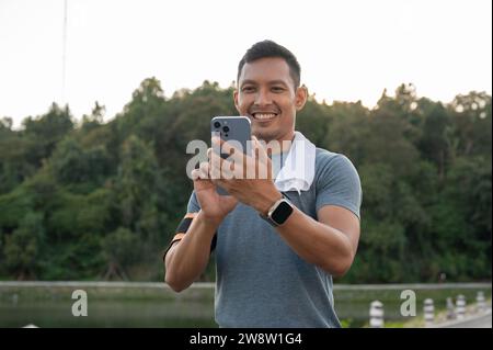 A handsome Asian man in sportswear is using his phone, reading messages or checking emails during his morning jogging in a park. lifestyle concept Stock Photo