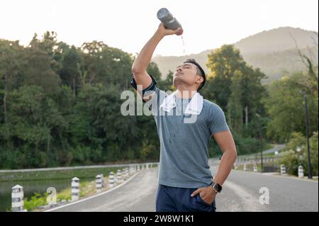 A thirsty, tired Asian man in sportswear is drinking pouring water from a bottle on his face after a long run outdoors in the morning. Stock Photo