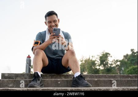 A happy and handsome Asian man in sportswear is sitting on the stairs and chatting with his friends on his phone after exercising running in a park. Stock Photo