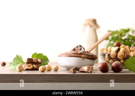 Chocolate cream with hazelnuts in a bowl and portions on wooden table with leaves and nuts around and white isolated background. Front view. Stock Photo