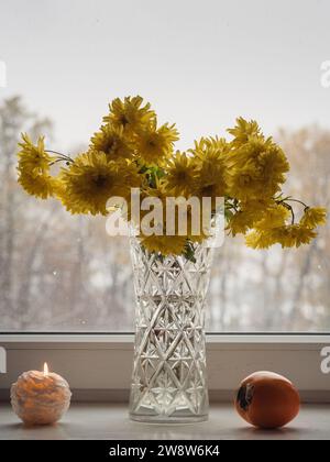Vertical table top view of yellow chrysanthemums in a glass vase, candle lit and a vibrant orange persimmon on a white with a winter forest panorama. Stock Photo