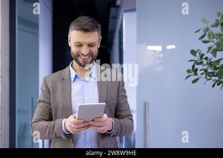 Mature experienced businessman inside office smiling contentedly, senior man holding tablet computer in hands, boss in business suit using analytical financial application. Stock Photo