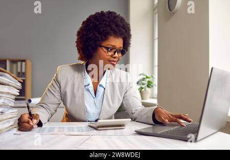 Young woman accountant working on a laptop at office with pile of folders and a stack of papers. Stock Photo