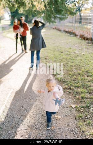 Little girl is standing on a path against the background of a photographer taking pictures of a married couple with a child Stock Photo