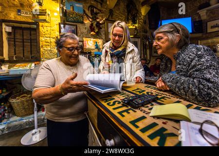 Restaurant owner Rosina Tabone (left) shows journalist Angela Berg (centre) and a guest (right) something in an open book at the counter of her restaurant Ta' Rosina in Sannat, Gozo, Malta. The dining room seats only a few tables and is popular with villagers and visitors from Malta alike for its traditionally cooked national dishes fenek (rabbit) and lampuki (dolphinfish). Stock Photo