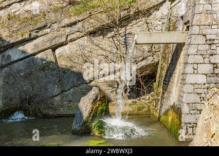 Crystal clear clean water gushing from fountain onto a stone with moss in a basin, wall of huge stones with wild plants between the grooves in the bac Stock Photo