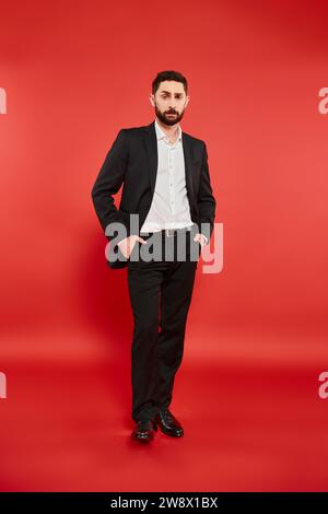 confident and stylish businessman in black suit standing with hands in pockets on red, full length Stock Photo