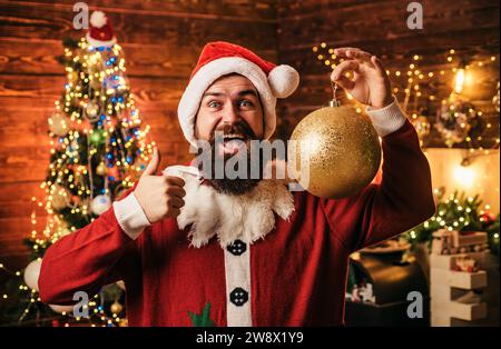 Crazy, funny Hipster Santa. Portrait of happy Santa with decorative toy balls near Christmas tree. Happy Santa dressed in winter clothing think about Stock Photo