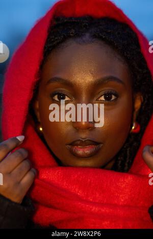 This portrait captures the intense and enigmatic gaze of a young African woman wrapped in a vibrant red scarf. The photograph is taken during the blue hour, which casts a serene hue, contrasting dramatically with the warm red of the scarf. Her piercing eyes are the focal point, highlighted by the subtle interplay of light and shadow on her face. Her complexion glows with an understated luminosity against the deep blue background, while her subtle makeup and simple jewelry add touches of elegance without distracting from the raw emotion of her expression. Enigmatic Gaze at blue hour. High quali Stock Photo