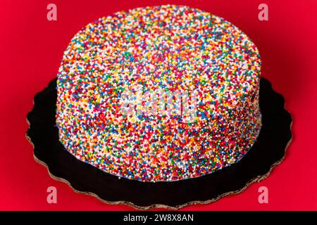 A cake with white icing encrusted in rainbow sprinkles on a red background. Stock Photo