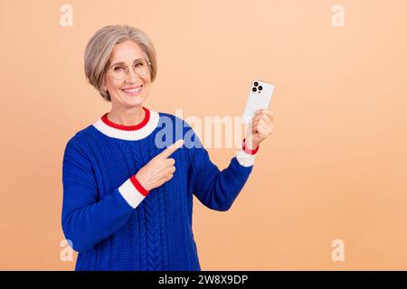 Photo of charming positive woman dressed trendy blue clothes presenting new iphone isolated on beige color background Stock Photo