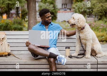 Smiling young man with laptop stroking dog while sitting on bench in park Stock Photo
