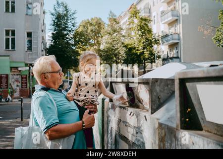 Senior man carrying granddaughter trashing garbage in can at street Stock Photo