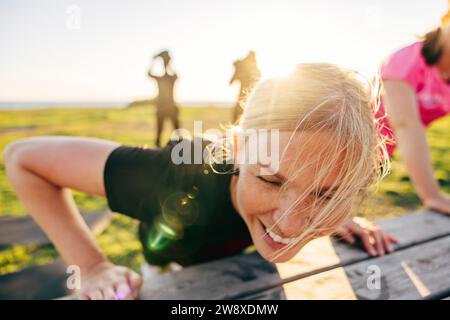 Smiling blond woman doing push-ups on bench during group training at beach Stock Photo