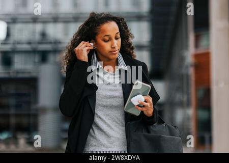 Young businesswoman wearing wireless in-ear headphones while holding smart phone Stock Photo