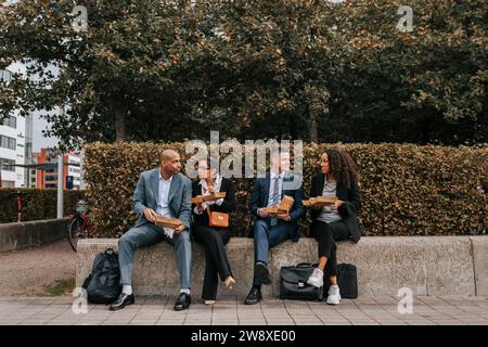 Male and female corporate colleagues having lunch while sitting on bench near plants at sidewalk Stock Photo